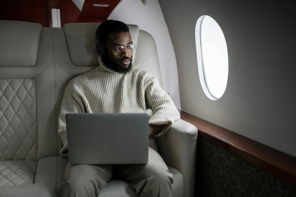 A man with glasses and a beard wearing a cream-colored sweater works on a laptop while seated in the luxurious quilted leather interior of a private jet, with natural light streaming through the circular window beside him.